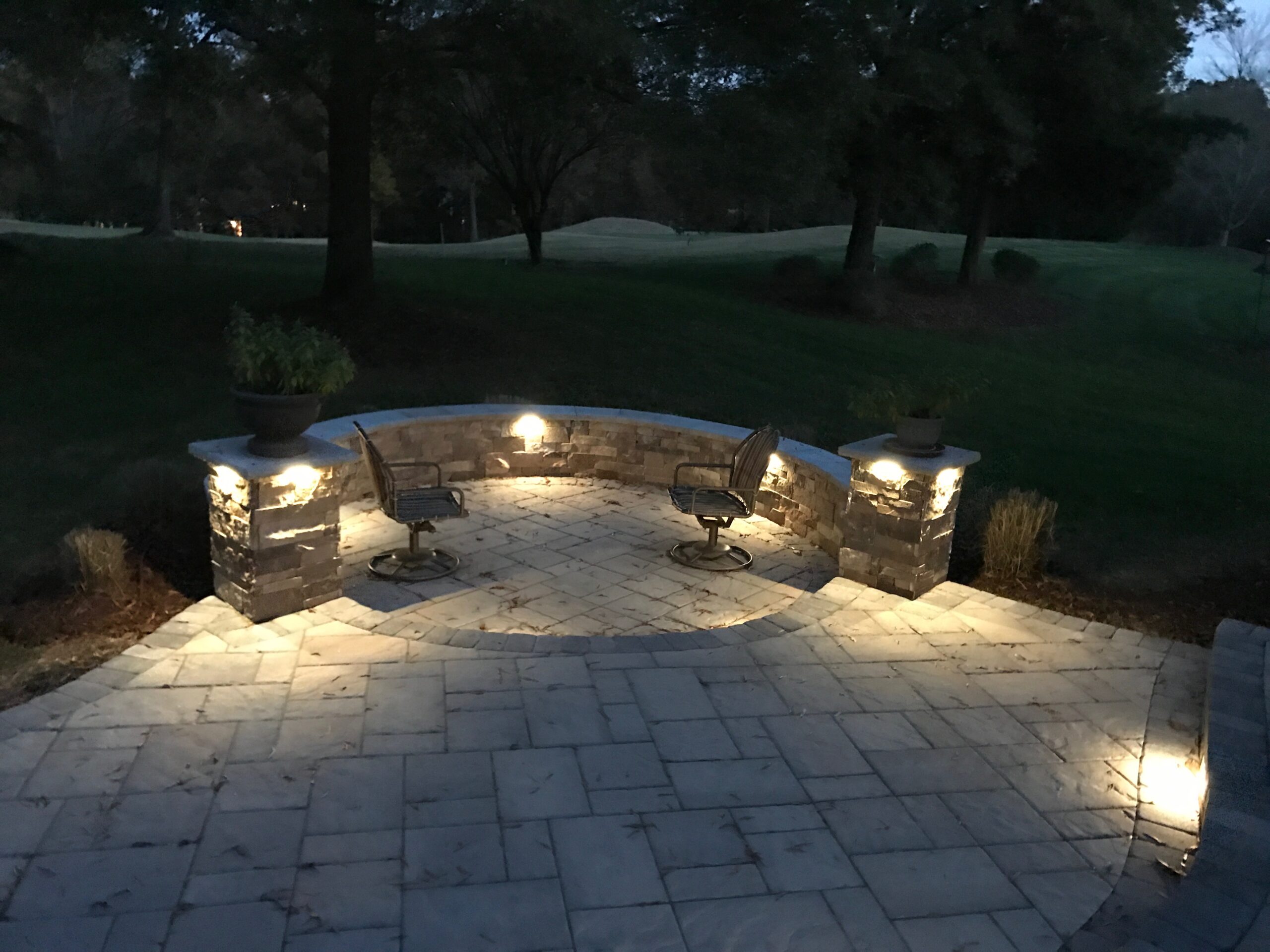 A beautiful and well done stone pavement with a stone sitting wall, embed outdoor lightning and chairs at the bottom of the photo.