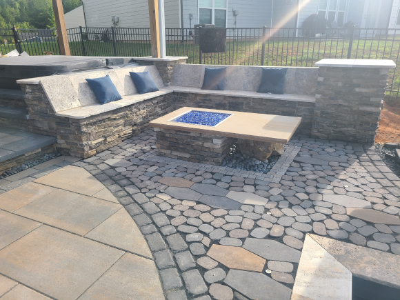A stone fire pit in a patio. There's a concrete L shape sofa with cushions on it. Seat walls.