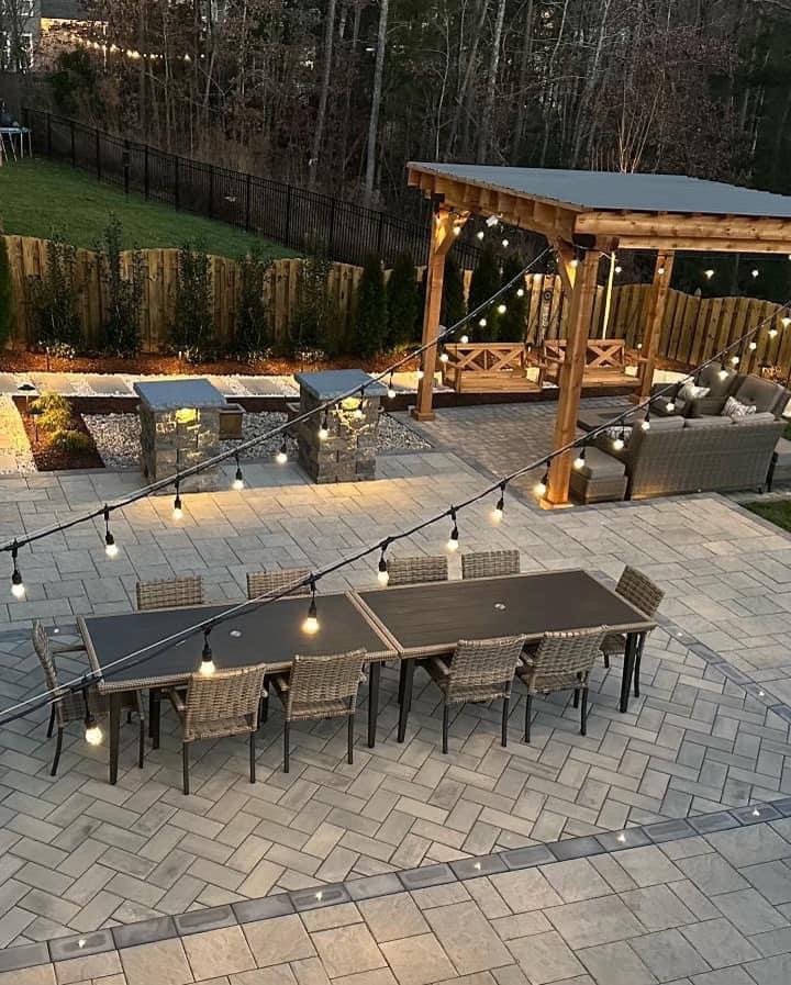 A patio where you can see a stone floor, tables for outdoor dining, a pergola and lights hanging from the wall of the house to the pergola. A perfect Social Space.