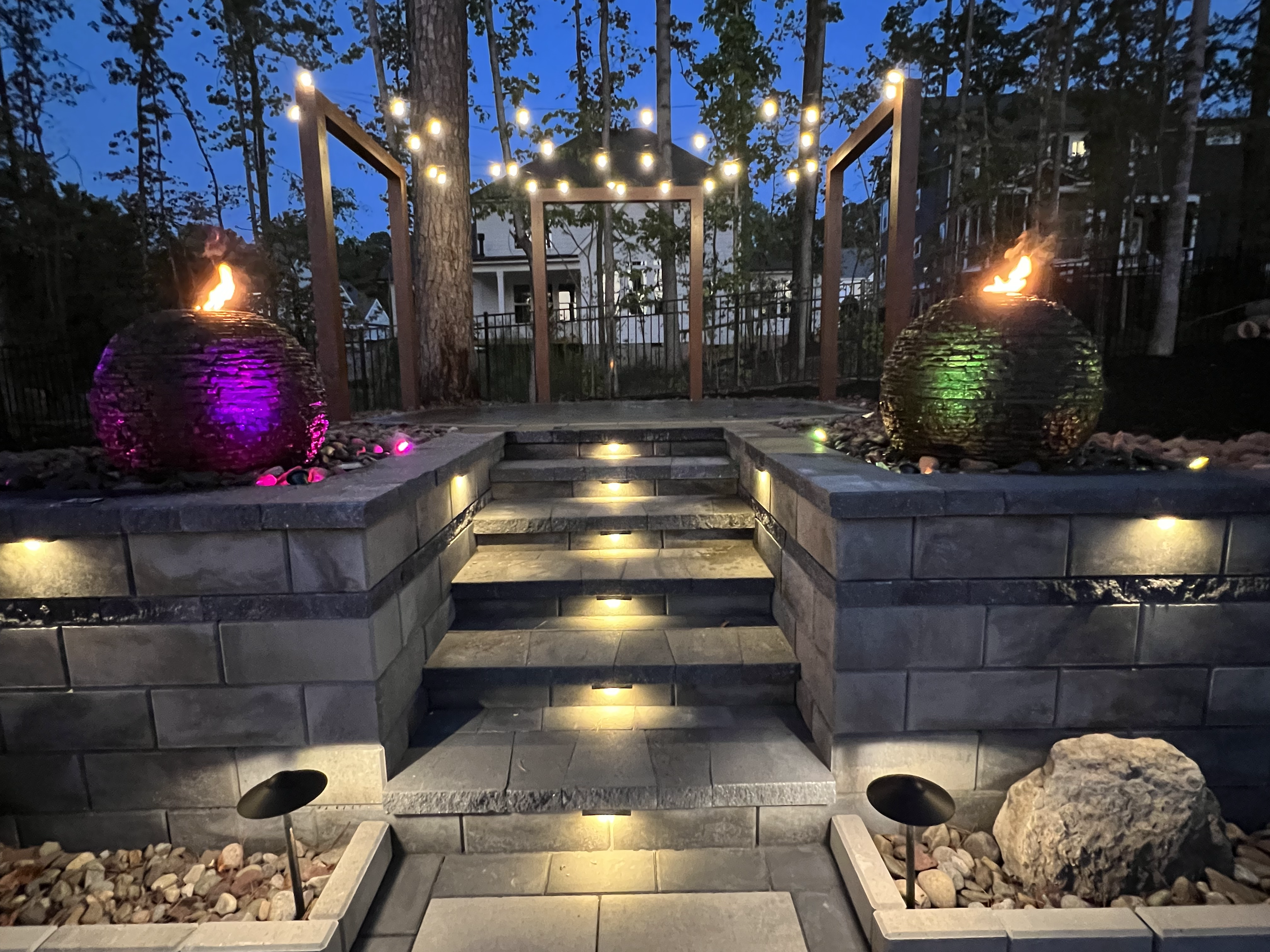 A stone patio with decorated with outdoor lightings such as hanging and embed ones. There's also 2 round rock fountains with fire effects