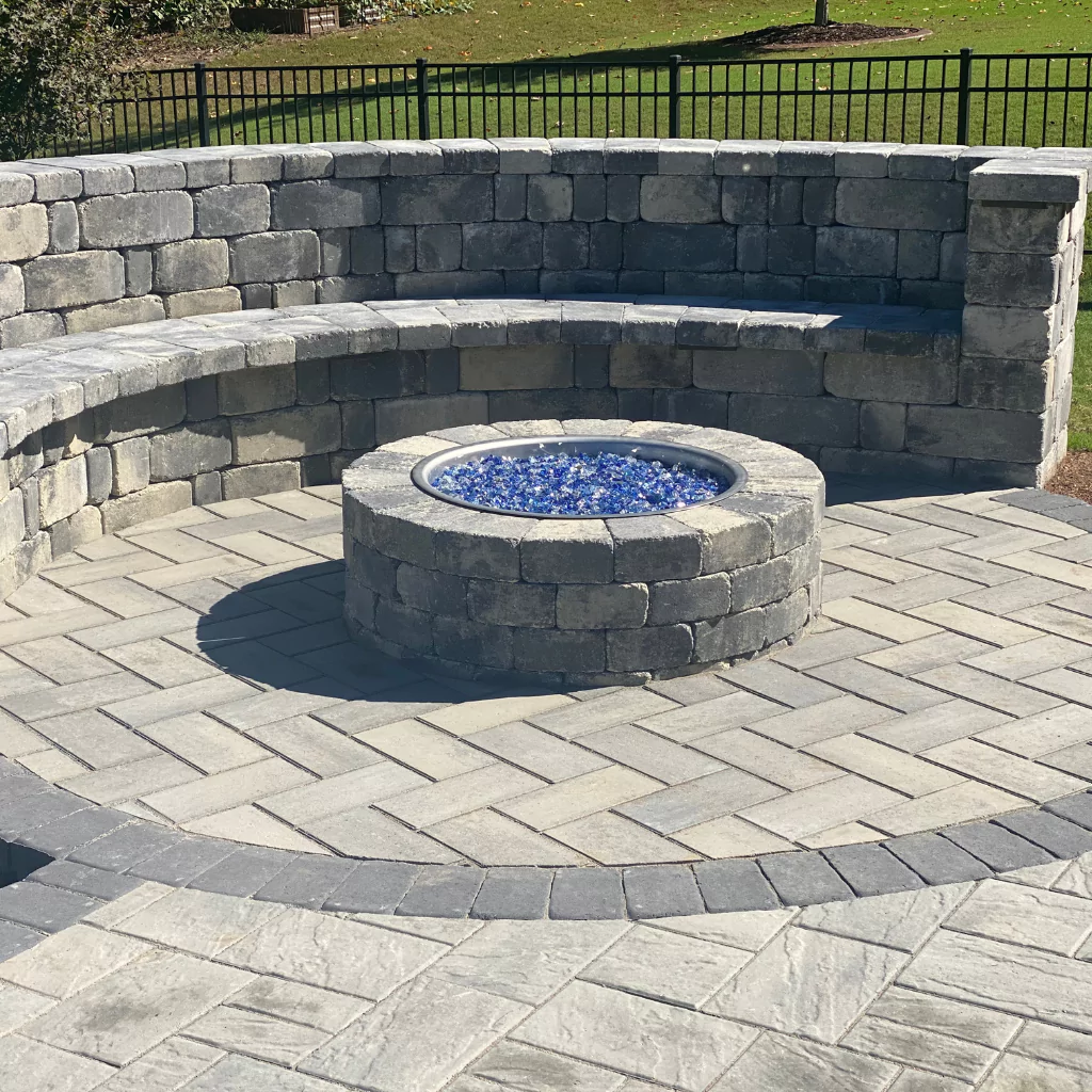 A stone fire pit with blue glass crystals inside. There is a stone wall to sit around it and also an elegant stone floor that makes the whole set combine.