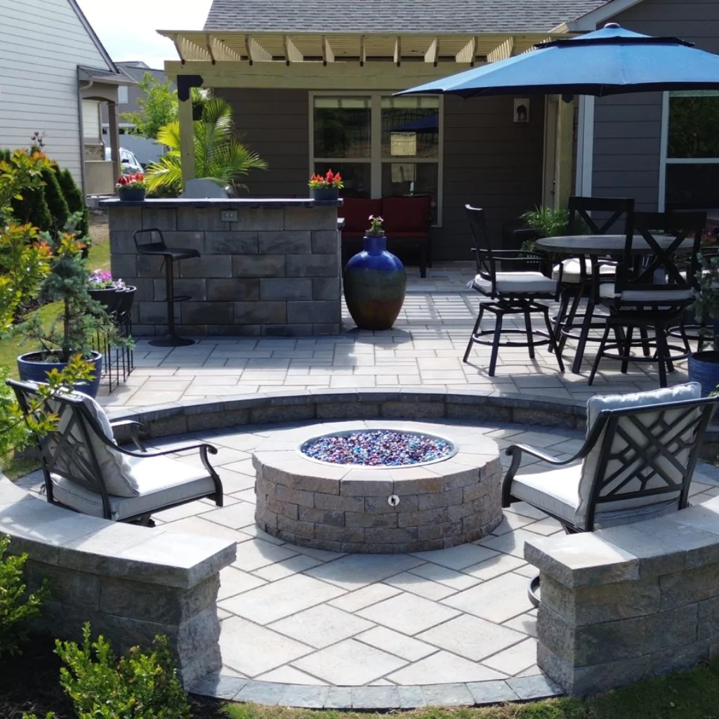 A patio with a fire pit surrounded by a semi-circular wall and metal chairs.