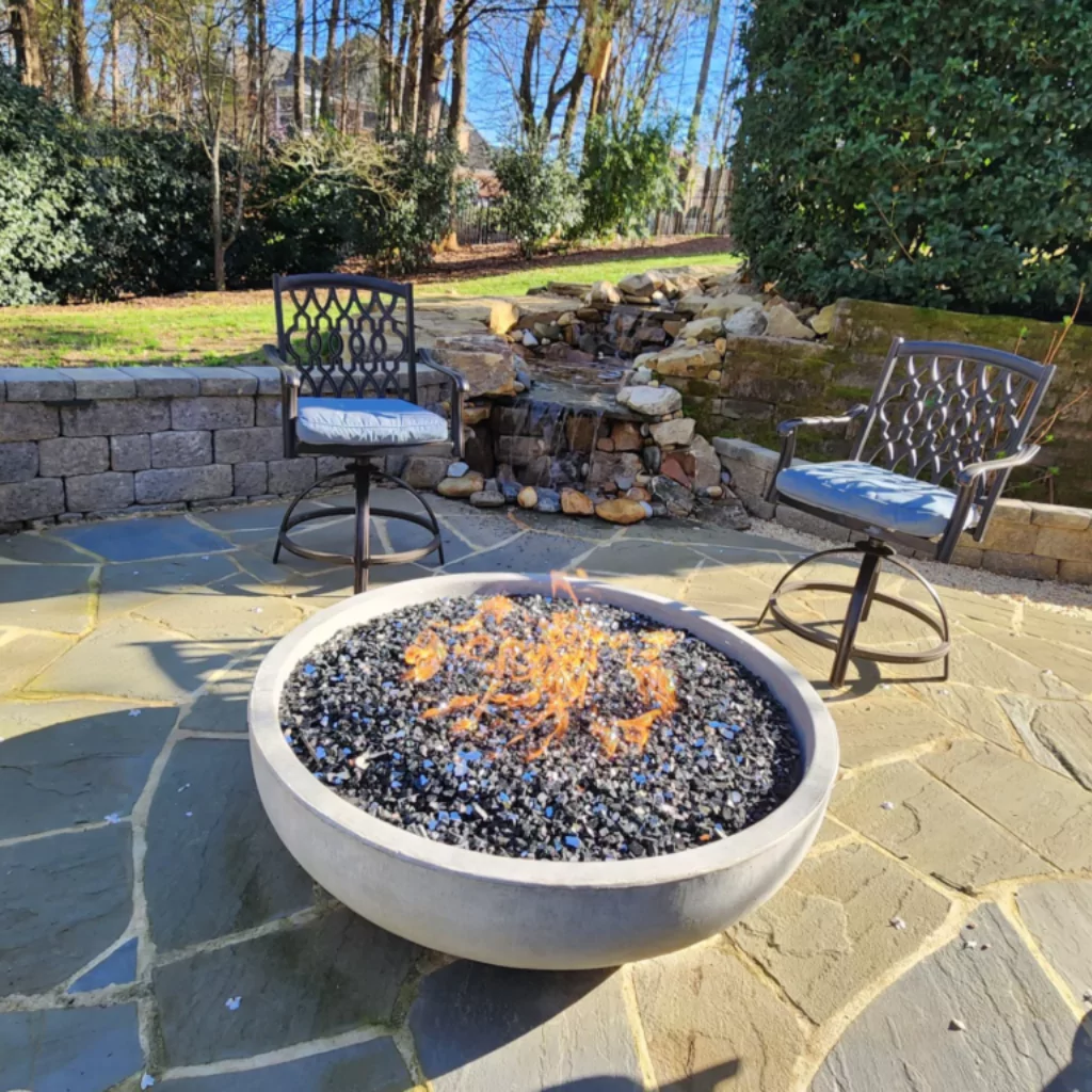 Fire Pit in a patio with metal chairs. There's a stone fountain and a sitting wall behind them.