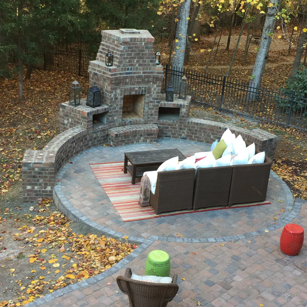 A semi-circular outdoor fireplace built in a beautiful gray stone. In front of it, there is a sofa and outdoor furniture.
