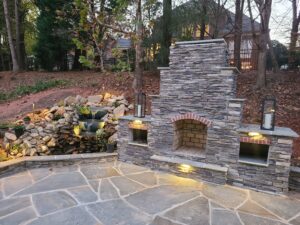 The natural beauty of a bluestone flagstone patio in Charlotte is enhanced by a custom stacked stone fireplace and a cascading boulder waterfall, all expertly lit by SunSouth Carolinas. Fall patio.