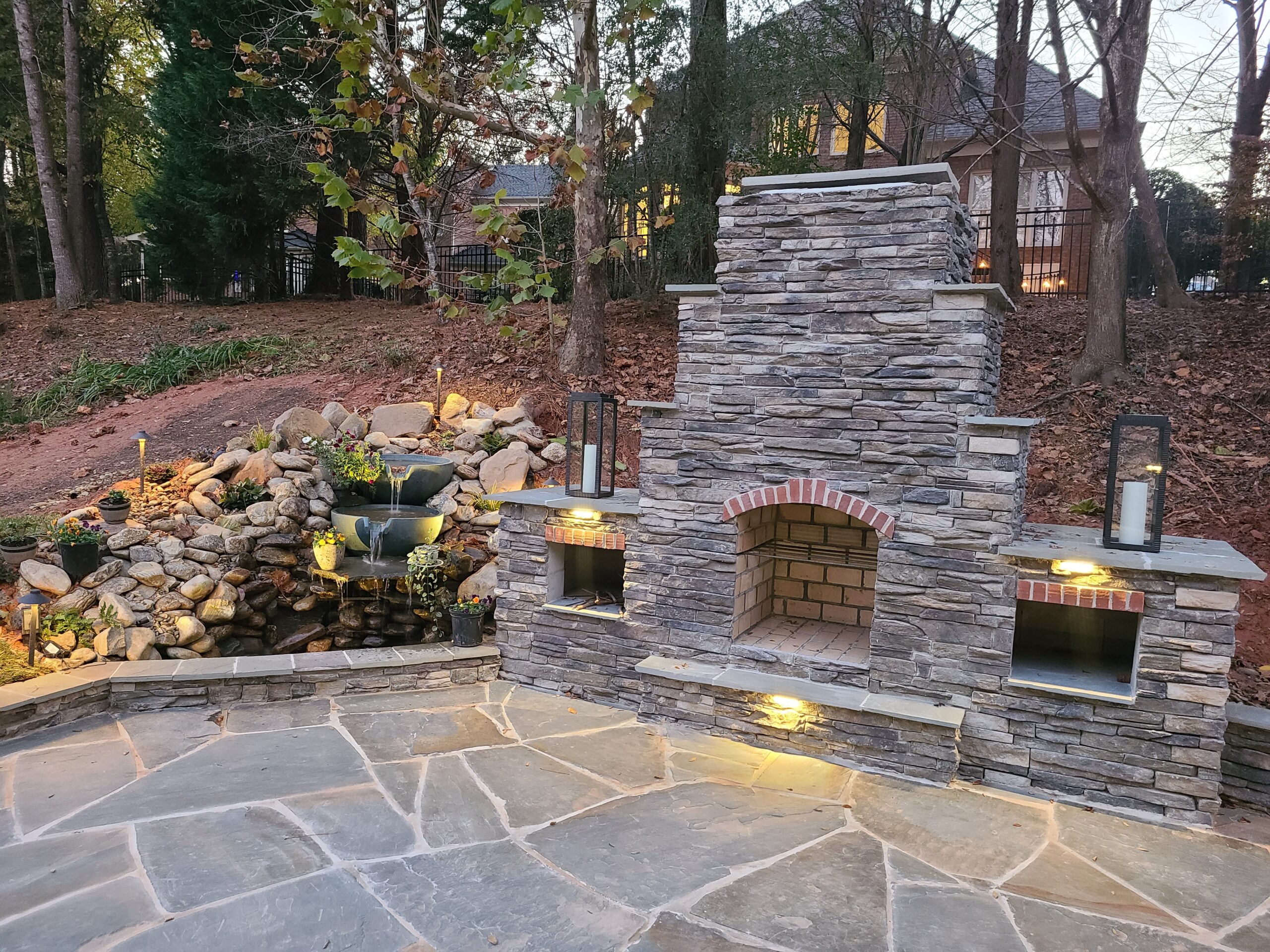The natural beauty of a bluestone flagstone patio in Charlotte is enhanced by a custom stacked stone fireplace and a cascading boulder waterfall, all expertly lit by SunSouth Carolinas.
