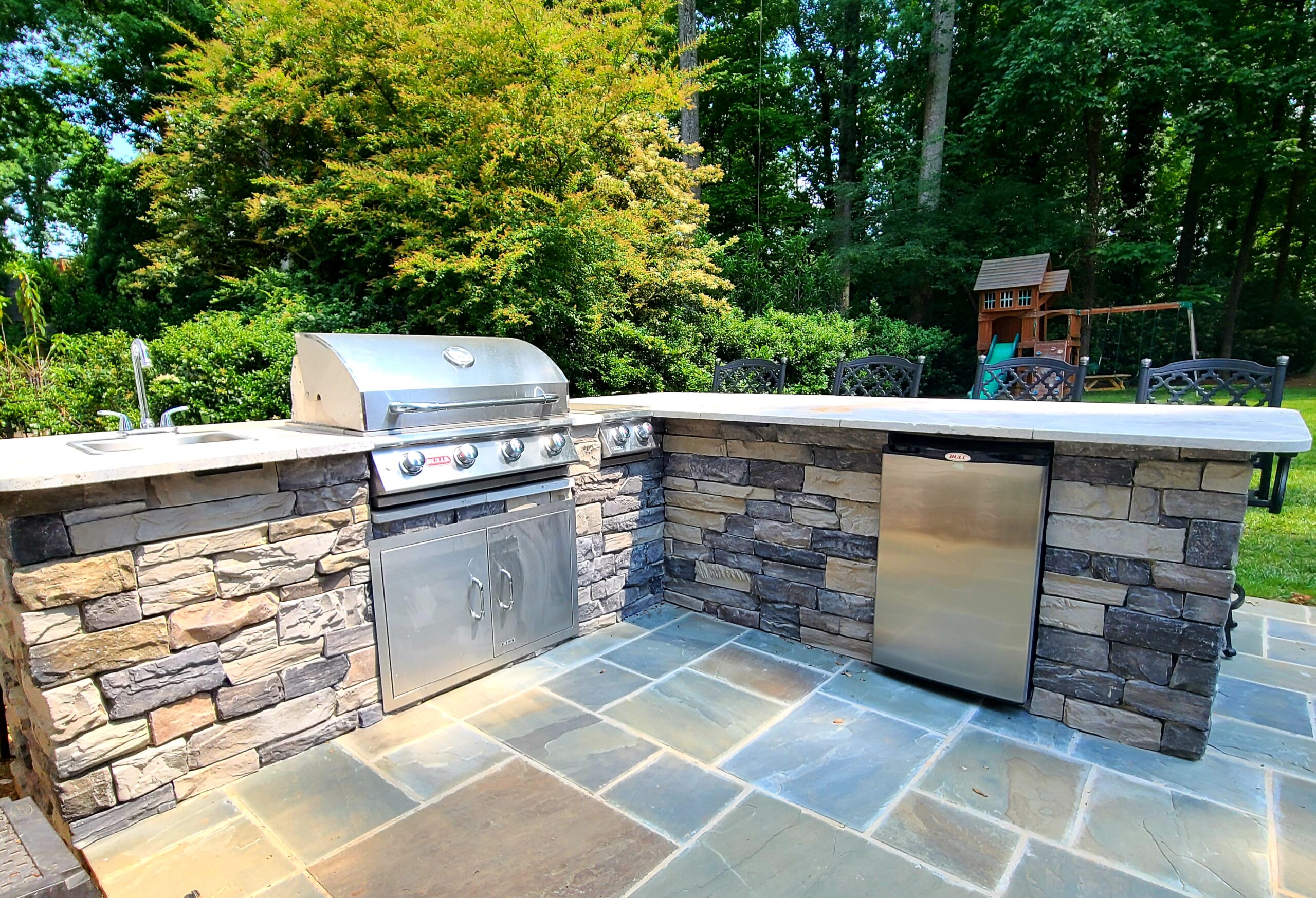 A metal outdoor kitchen with a little fridge, embeded in a stone deck. A lot materials blend in this photo.