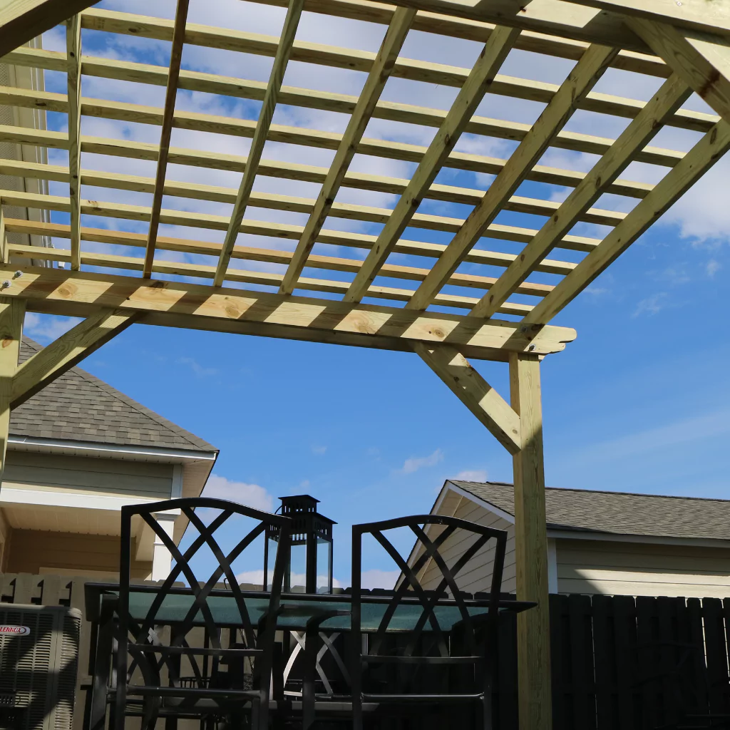 A pergola decorating a small patio. Metal chairs and an outdoor kitchen can be seen.