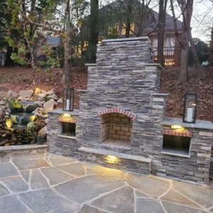 A stone fireplace in a stone-floored courtyard with a fountain on one side and a forest in the background.