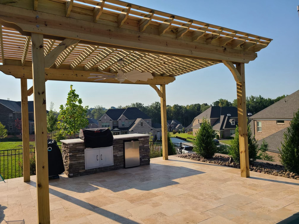 A pergola in a patio. The floor is made of stone and under the pergola is an outdoor kitchen.