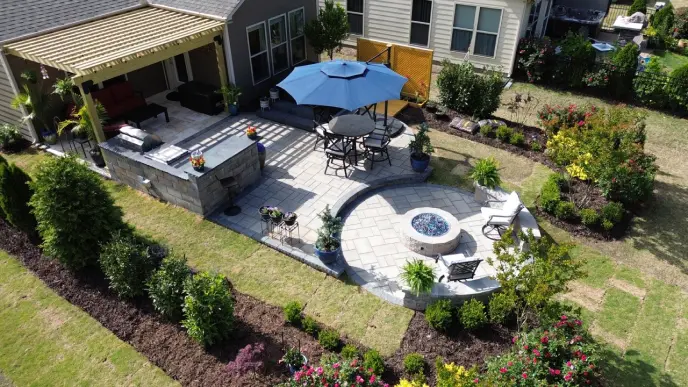 A patio shown from an aerial view. You can see a rock firepit in the center and a wall with some metal chairs around it. There is also a pergola and an outdoor kitchen.