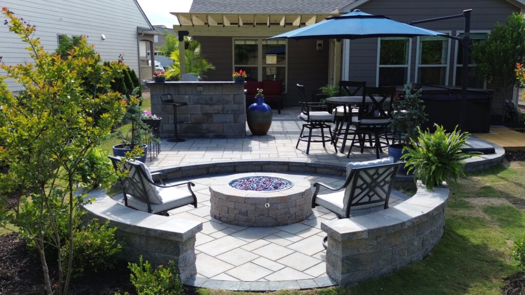 A sky view of a patio with a fire pit surrounded by a semi-circular wall and metal chairs. At the botom there's a pergola and an outdoor kitchen. Outdoor experience.