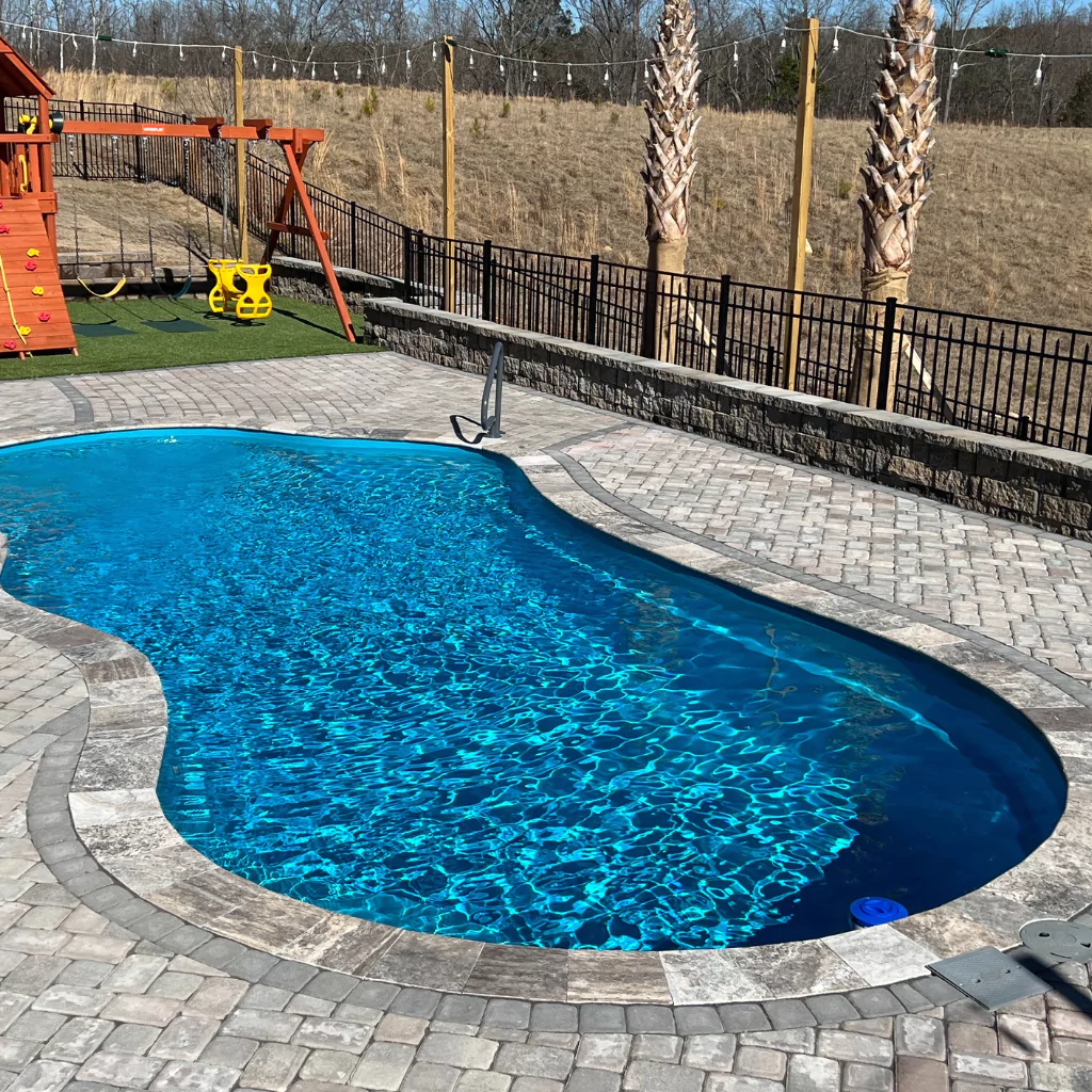 A swimming pool in a patio. It has a stone floor around it and a children's playground at the bottom.