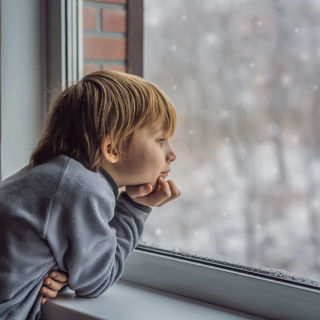 A child looking out of the window into his backyard. It's winter outside and there is a lot of snow