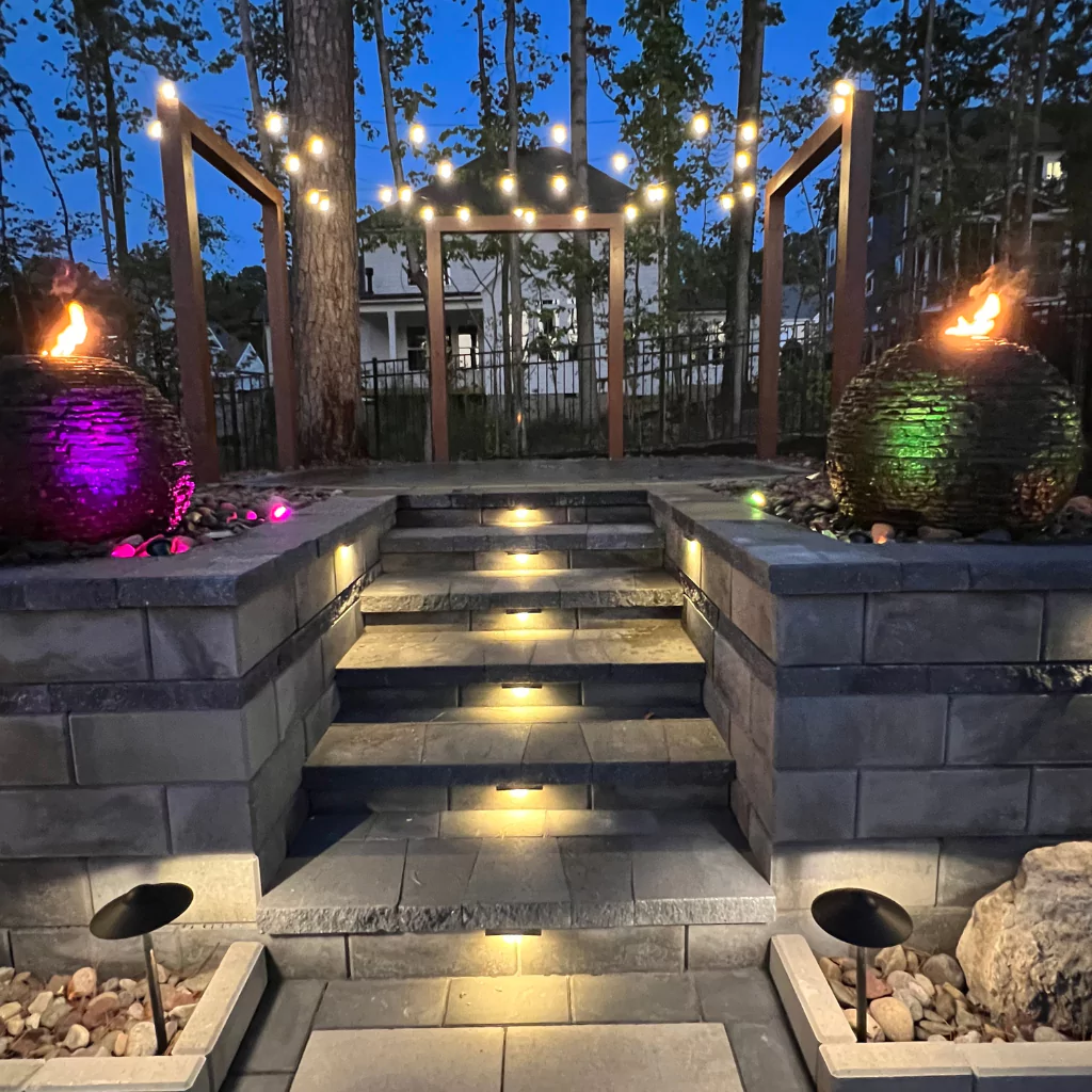 A stone patio with decorated with outdoor lightings such as hanging and embed ones. There's also 2 round rock fountains with fire effects