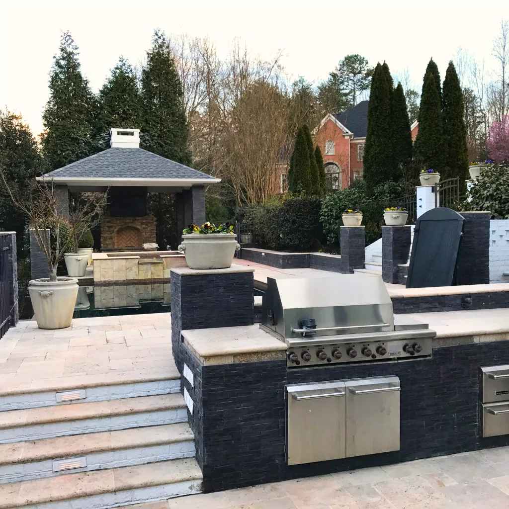 An elegant outdoor kitchen on a patio with stone details. The shiny stainless steel finish of the grill and refrigerator contrast nicely with the black stone and light-colored granite countertop.