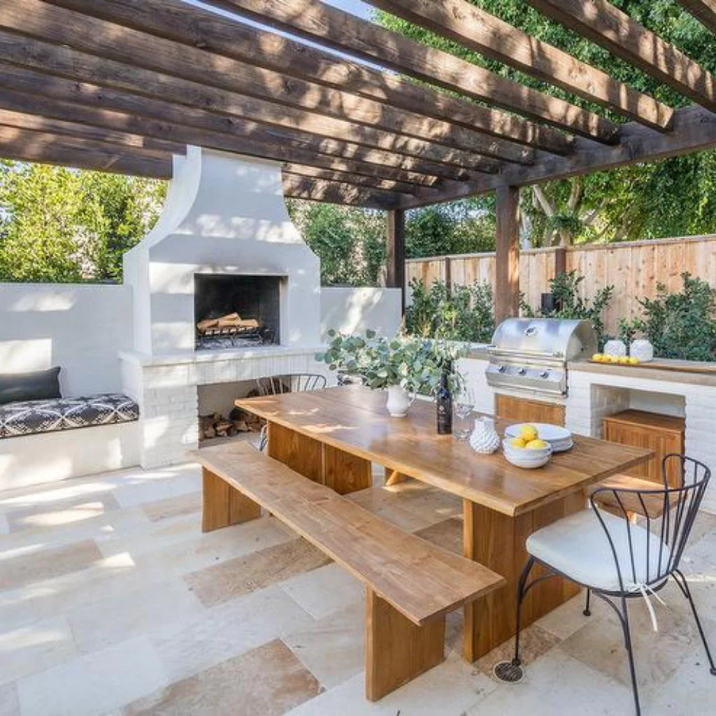 A dining table under a pergola. There's a fireplace and an outdoor kitchen at the bottom.
