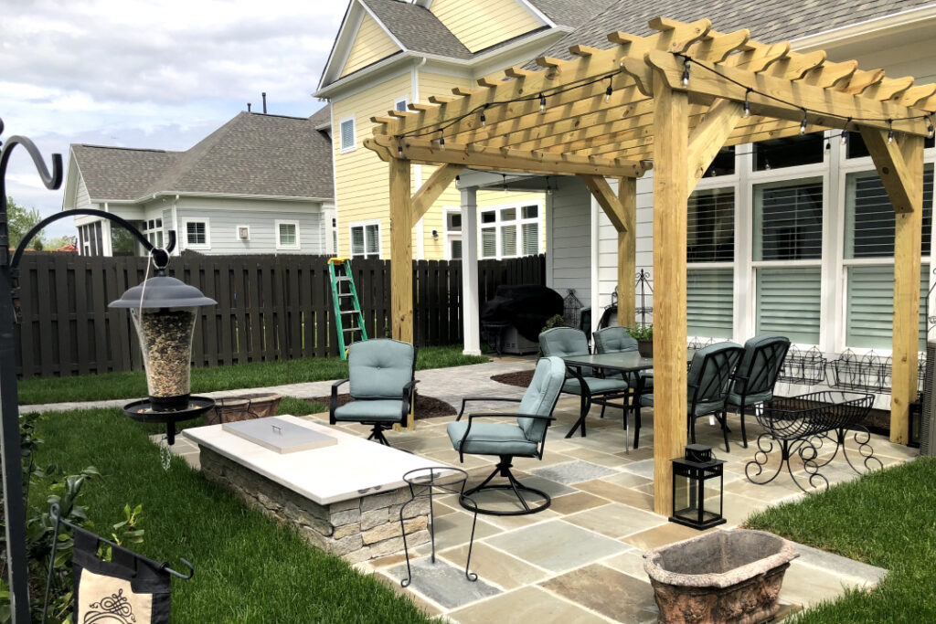 A pergola decorating a small patio. Metal chairs and an outdoor kitchen can be seen.