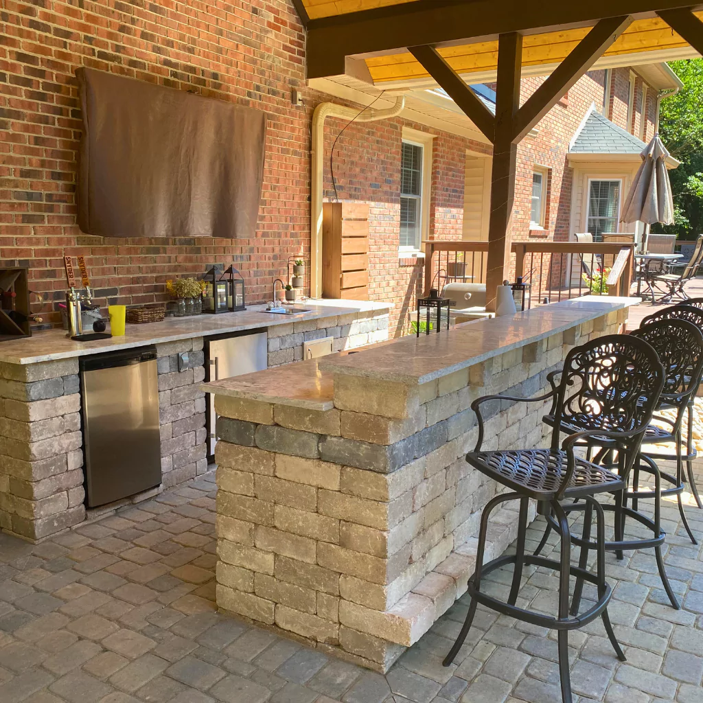 An elegant outdoor kitchen, full equipped with home appliances, a grill and chairs. It's under a wooden roofing.