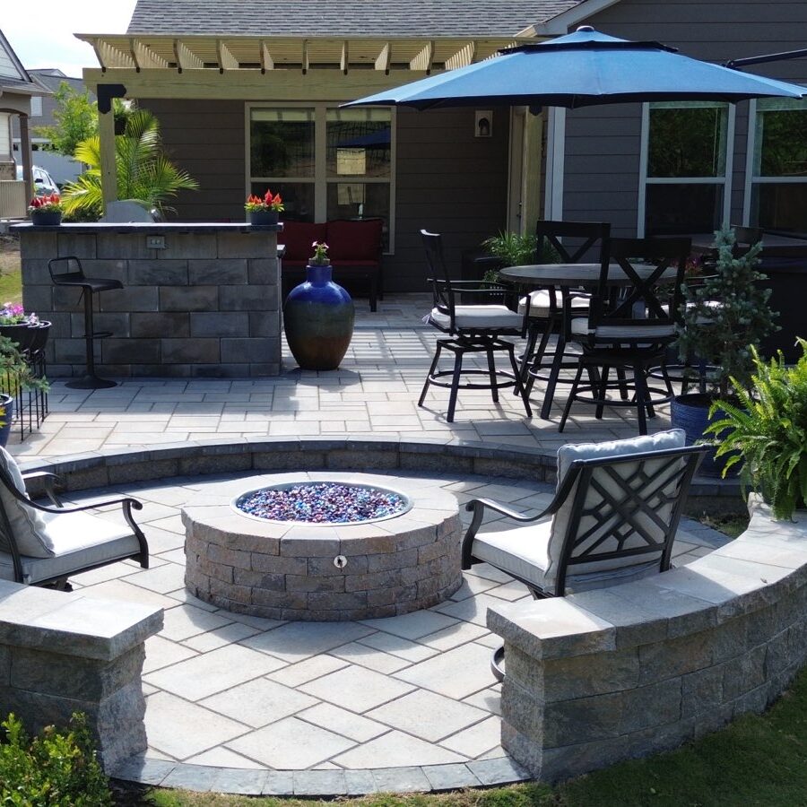 A sky view of a patio with a fire pit surrounded by a semi-circular wall and metal chairs. At the botom there's a pergola and an outdoor kitchen.