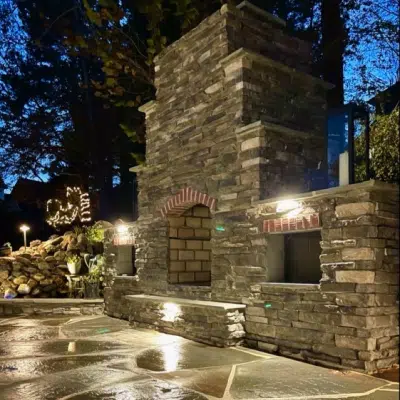 Romantic Outdoor Spaces: Fire Features Designed for Cozy Winter Nights 6 A stone fireplace in a stone-floored patio with a fountain on one side and a forest in the background.