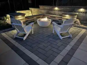 Modern outdoor living space by SunSouth Carolinas featuring a two-tone paver patio in herringbone pattern, sleek fire bowl, and multi-level retaining wall with built-in bench and lighting.