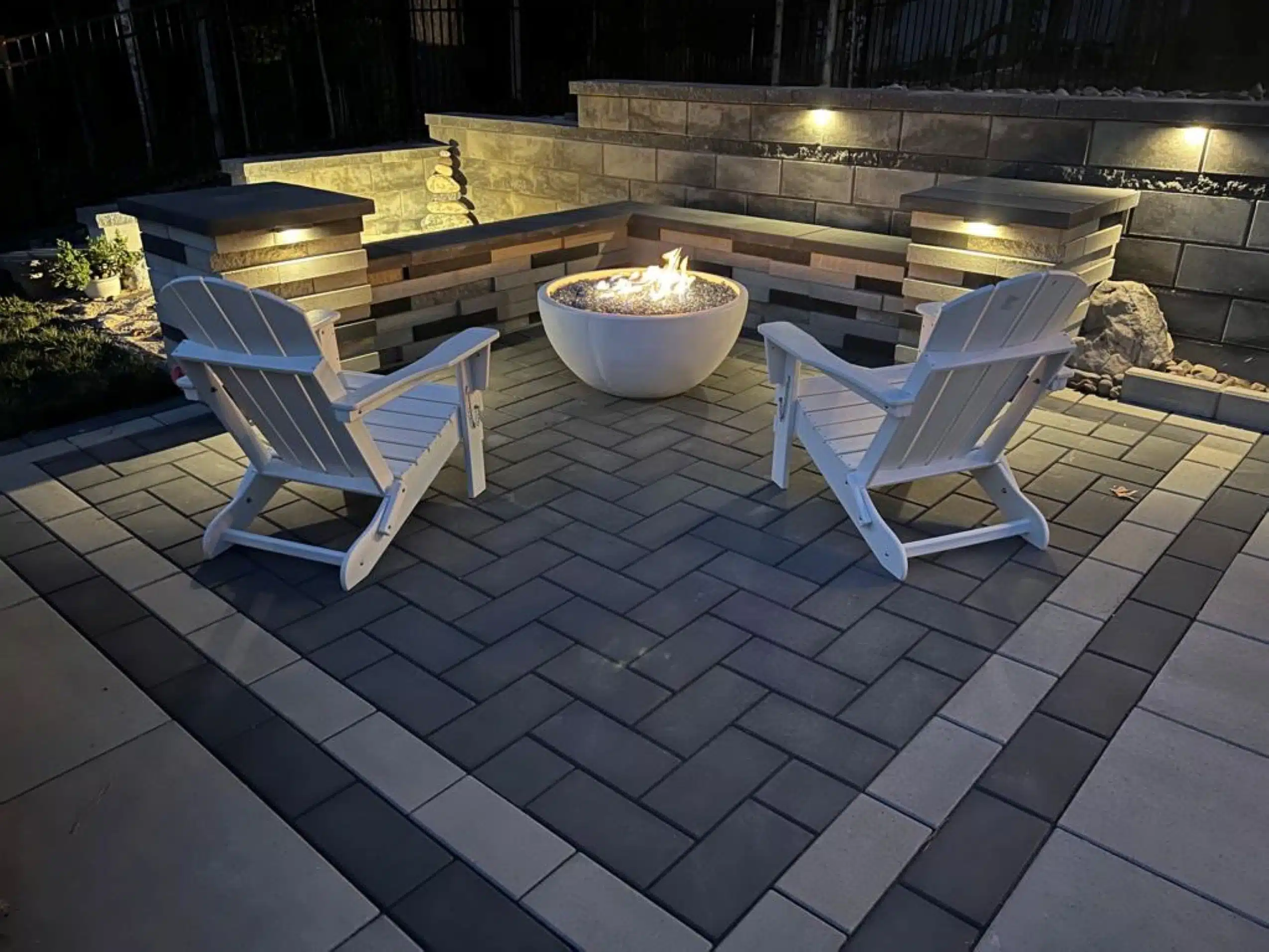 Gas vs. Wood-Burning Fire Pits: Pros, Cons, and Installation Costs 1 Modern outdoor living space by SunSouth Carolinas featuring a two-tone paver patio in herringbone pattern, sleek fire bowl, and multi-level retaining wall with built-in bench and lighting.