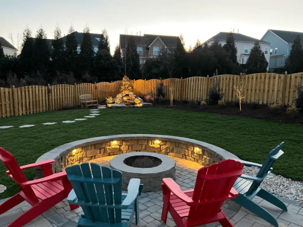 Cozy atmosphere created by SunSouth Carolinas with a lighted stone seating wall surrounding a fire pit, paver patio, and a distant illuminated water feature.