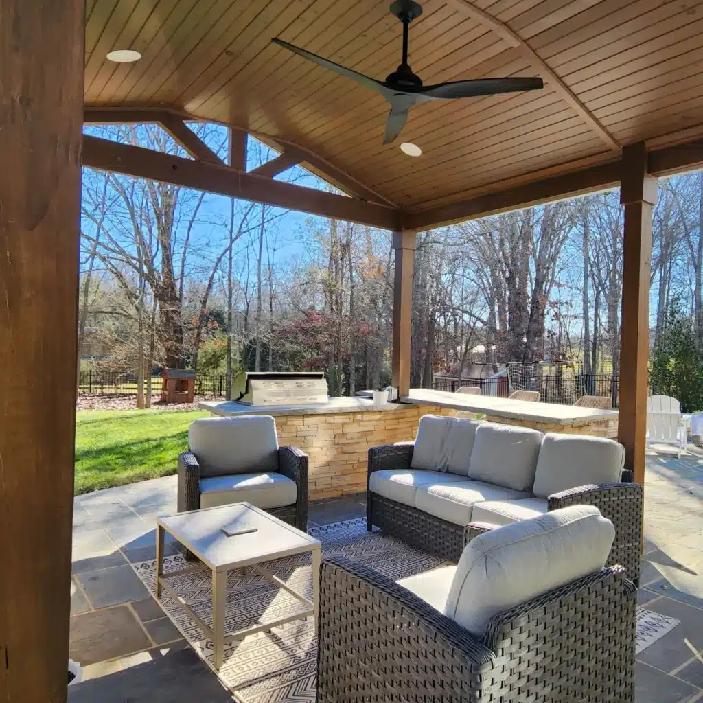 Luxury covered outdoor living space by SunSouth Carolinas featuring detailed masonry pillars, hardscaping floor, ceiling fan, recessed custom outdoor lighting, and custom outdoor kitchen.