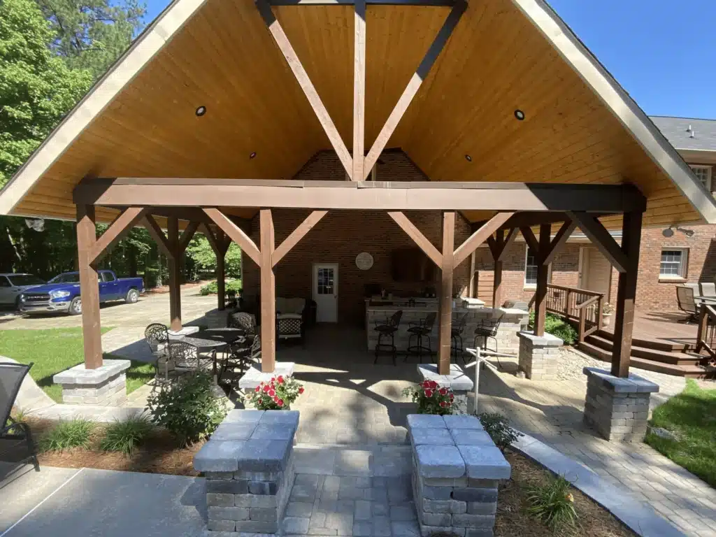 Grand outdoor pavilion designed by SunSouth Carolinas, featuring high wood-clad ceilings and trusses similar to custom wood pergolas, supported by masonry pillars on extensive hardscaping.
