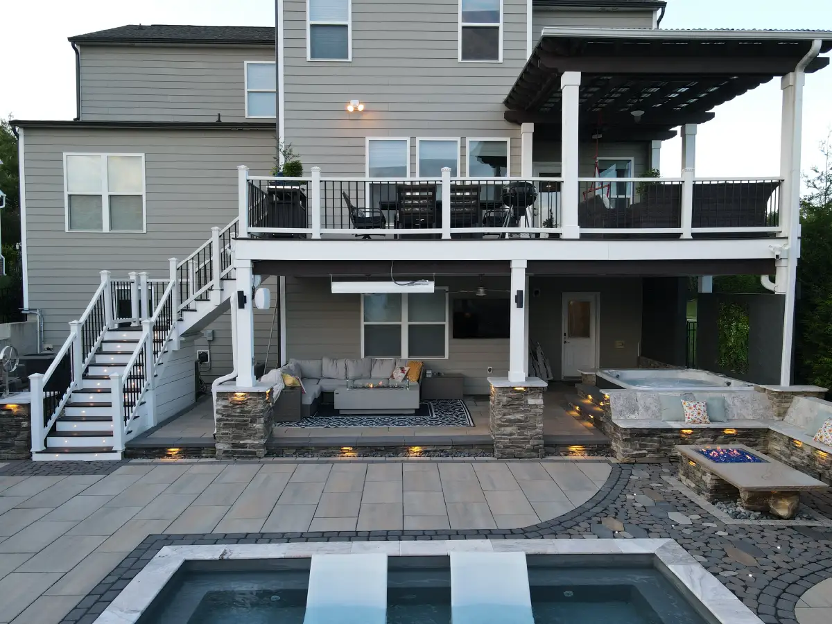 A multi-level outdoor living masterpiece where an elevated deck with a lighted staircase seamlessly connects to a covered lower patio, hot tub, and pool area by SunSouth Carolinas.