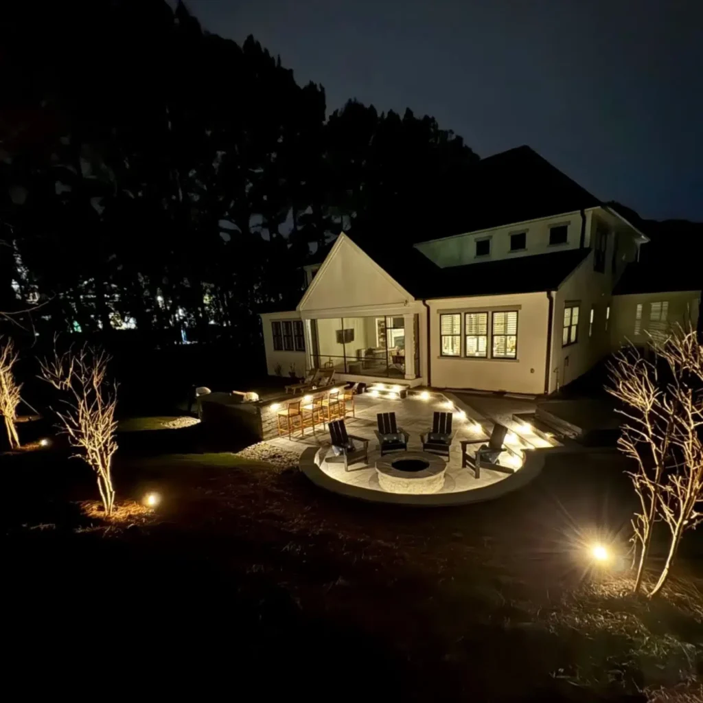 Romantic Outdoor Spaces: Fire Features Designed for Cozy Winter Nights 4 Multi-zone backyard design by SunSouth Carolinas illuminated by custom outdoor lighting service, showcasing hardscaped patio with stone fire pit and upper level with masonry seating walls.
